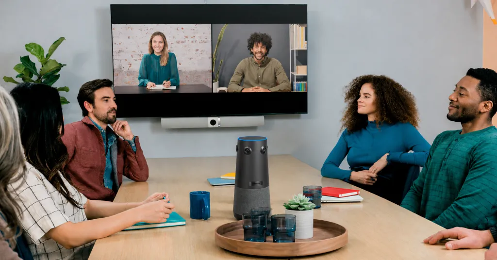 モダンな会議室の中央に360度カメラが設置されている様子。円形テーブルを囲んで参加者が座っており、全員の顔が正面から捉えられている。画面上の遠隔参加者も全員の顔を見渡せて、活発な議論が行われている。