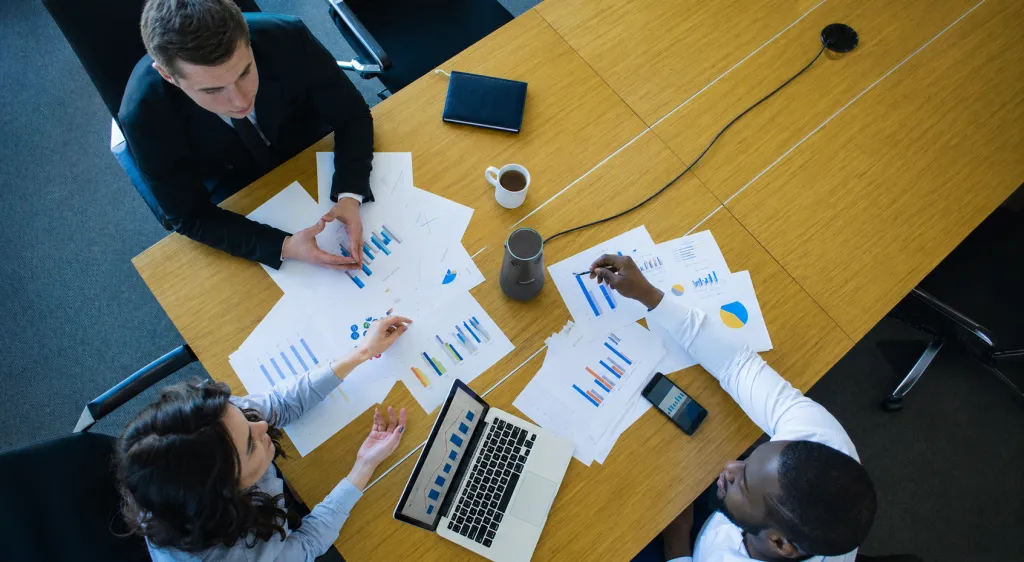 360Alien conference camera placed at the center of a consultation meeting table, connected to power while professionals review business charts, reports, and laptop data from an overhead view.