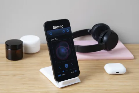 High-angle view of a modern desk setup featuring a smartphone playing music on a charging stand, over-ear headphones in the background, and a wireless earbud case on the table, illustrating a clean workspace ideal for the best wireless earbuds for Android