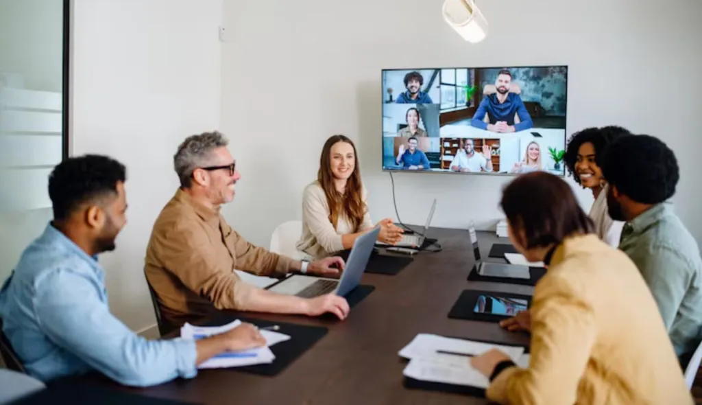 People participating in a hybrid meeting with a conference camera and AI tools displayed above the meeting table.