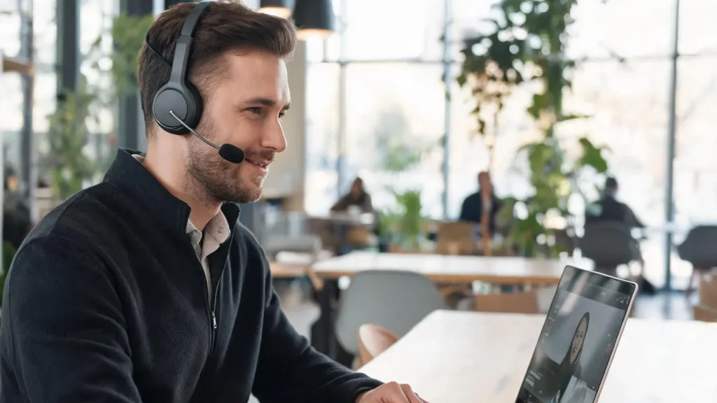A professional man with North American features wearing a dark grey wireless business headset while on a video call from a bright modern co-working cafe, laptop on a wooden table, blurred greenery background, relaxed confident posture.