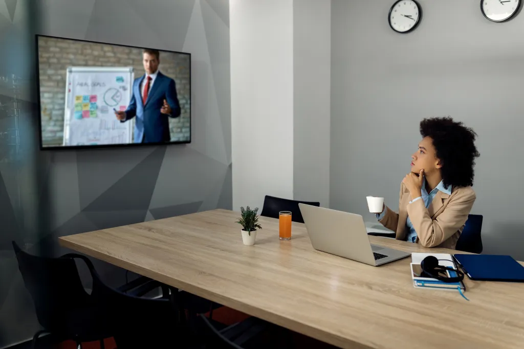 A woman in a small meeting room presenting to colleagues during a hybrid video conference using a wide-angle webcam.