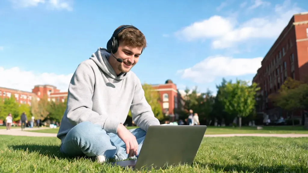 Student sitting on a grassy campus quad wearing a wireless headset, laptop open on the grass, sunny afternoon, relaxed candid lifestyle shot, natural outdoor lighting
