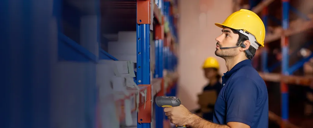 Inventory manager using a handheld scanner and wearing an open-ear wireless headset to manage stock in a high-density warehouse racking system.