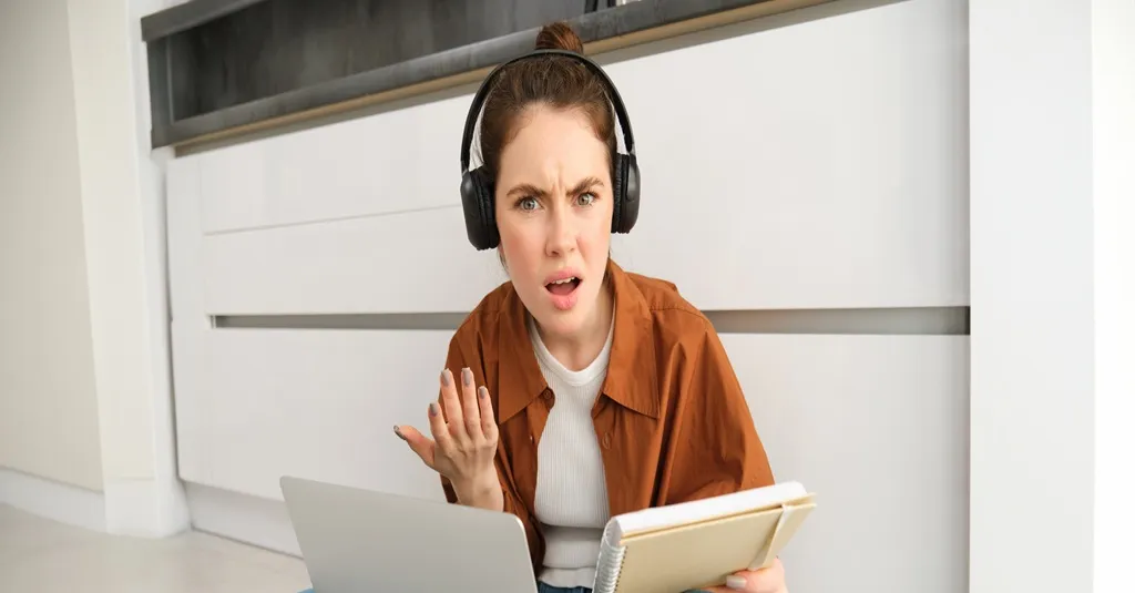 Woman feeling confused and annoyed wearing over-ear headphones while working on a laptop
