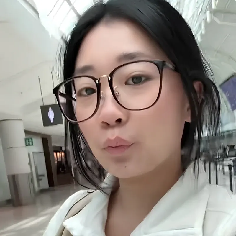 A woman wearing black round glasses taking a selfie in a bright indoor setting, showcasing open-ear awareness in daily life.