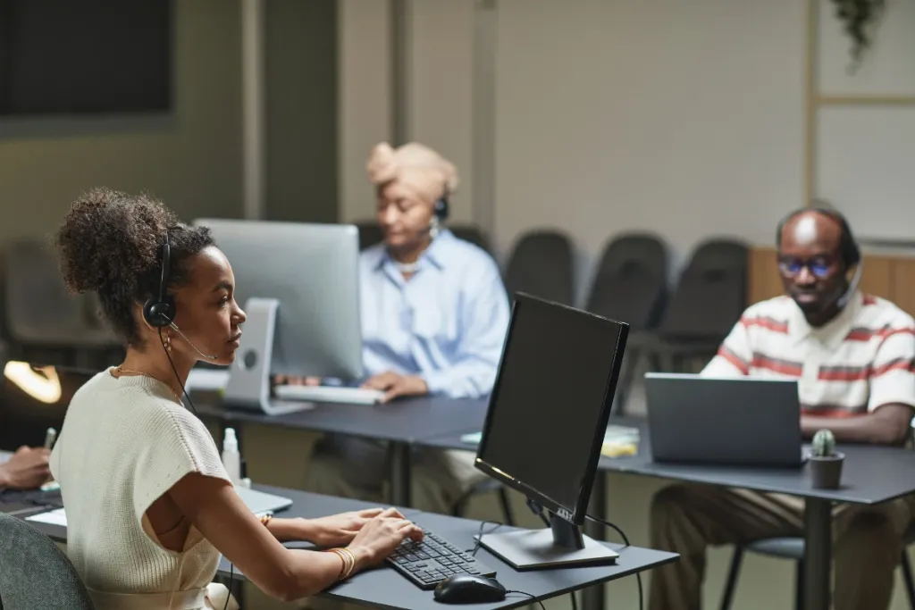 Line of call center representatives wearing headsets and sitting at desks in front of a large city-view window.