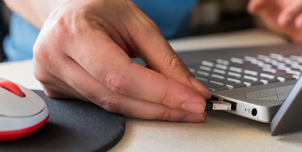 Close-up of a person plugging a black USB cable into the port of a laptop beside a computer mouse