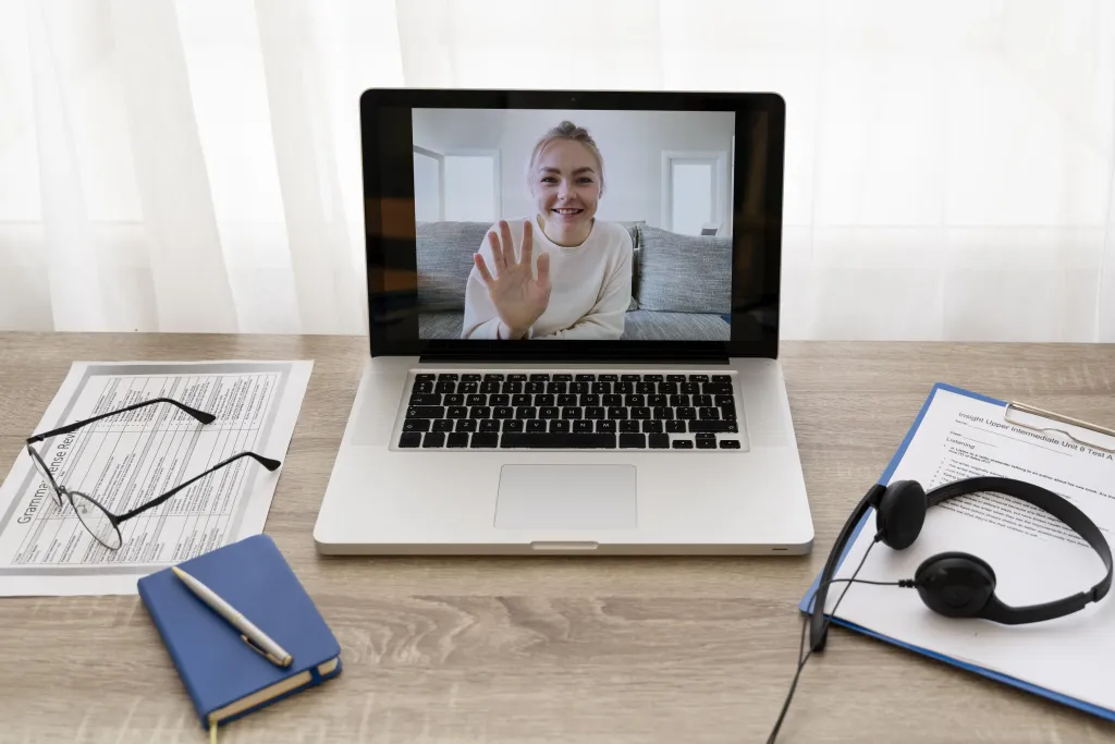 A student waving during an online class on a laptop setup with essential online teaching tools, a headset, and lesson materials on the desk.