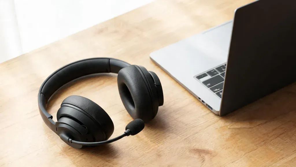 A wireless headset with a boom mic resting on a wooden desk next to a laptop, an authentic product shot with soft window light