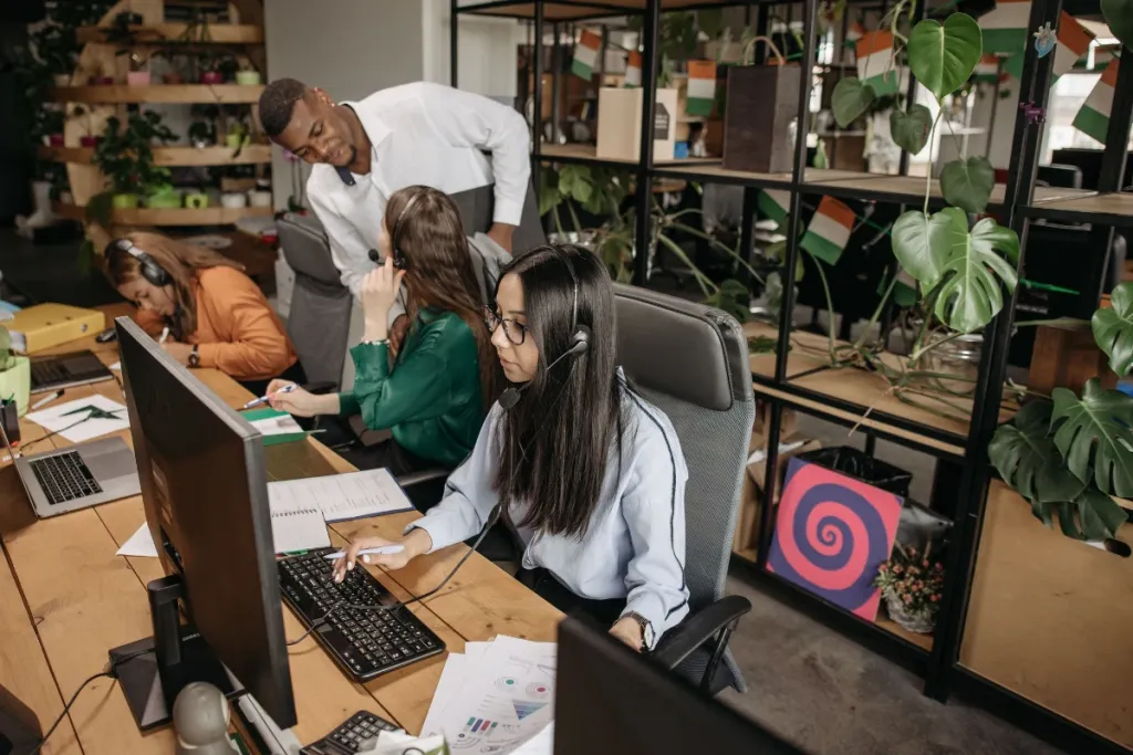 Male and female call center representatives wearing wireless headsets and checking document paperwork while working at computer desks.