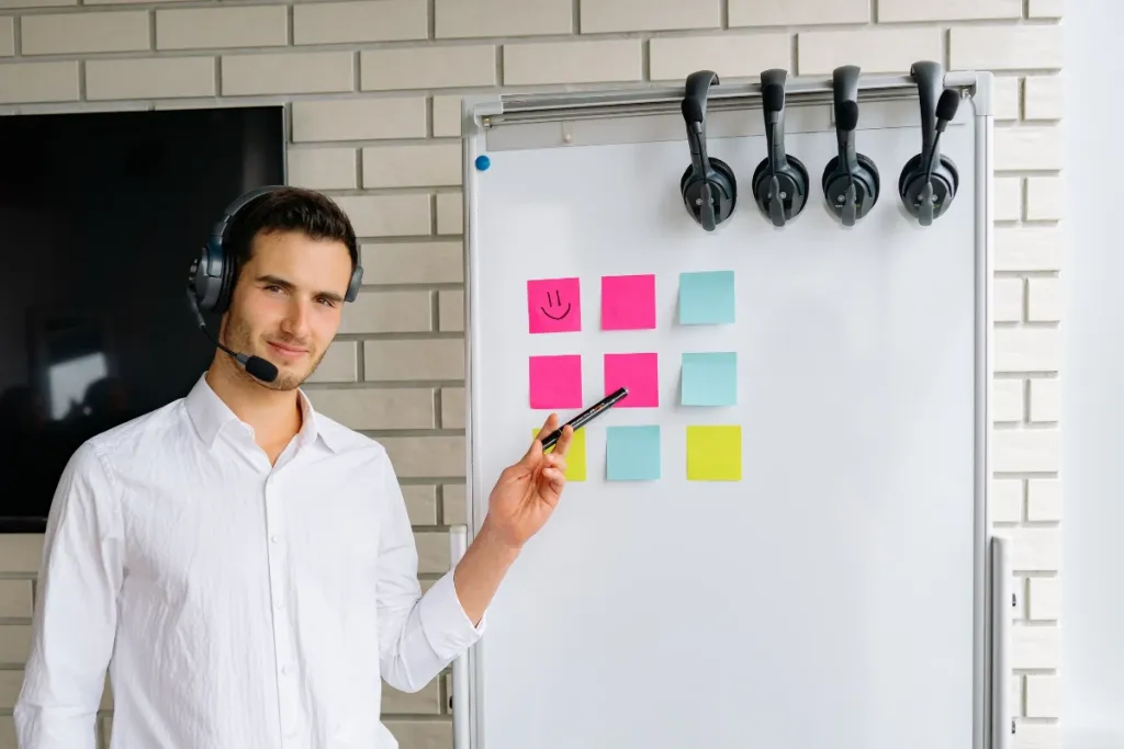 A man wearing a headset with a microphone points a marker at a whiteboard with colorful sticky notes and four more headsets hanging.