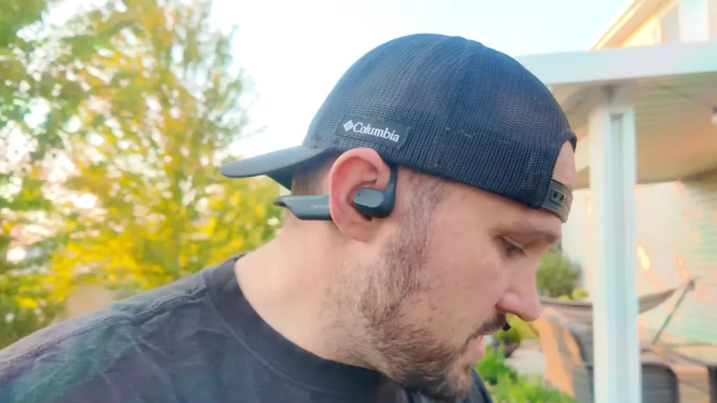 Side view of a man wearing a backward cap and black Nuroum open-ear headphones outdoors.
