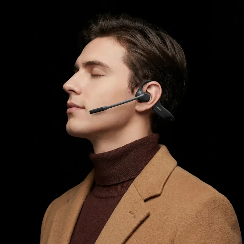 Side profile of a man wearing a brown coat and black Nuroum open-ear headset with a boom microphone against a dark background.