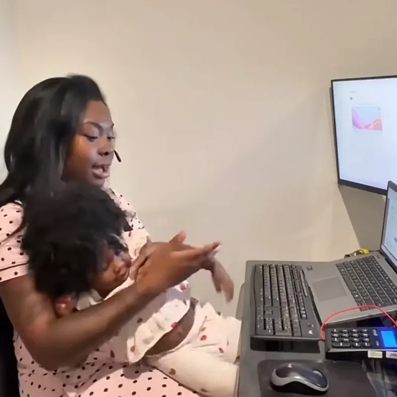 A woman multitasking at her desk, holding her toddler while wearing a Nuroum open-ear wireless headset.