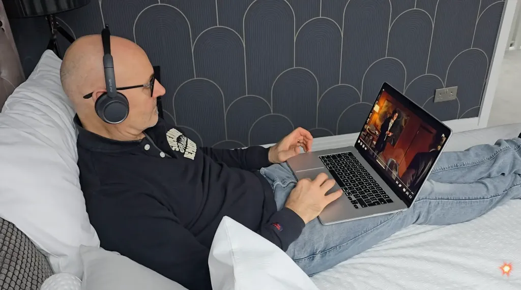 Tim wearing the Nuroum HP31D wireless headset with mic while watching videos in bed, showcasing its over-ear comfort and immersive sound for media playback