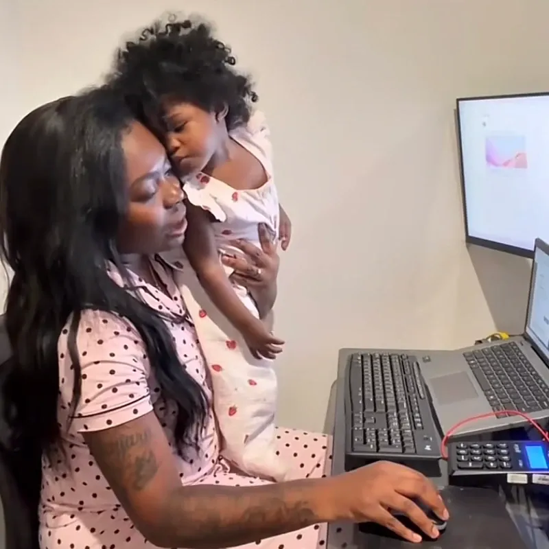 A mother working from home at her computer desk while comforting her toddler on her lap, showcasing real-life remote work challenges.