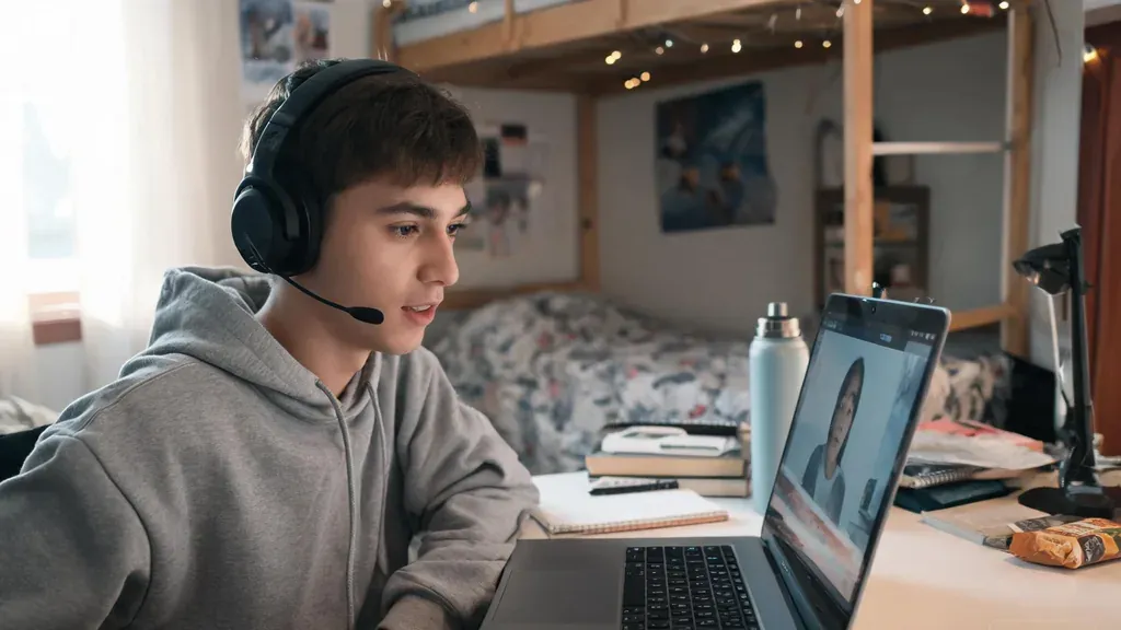 A student wearing a wireless headset with a boom mic while attending an online class on a laptop in a dorm room