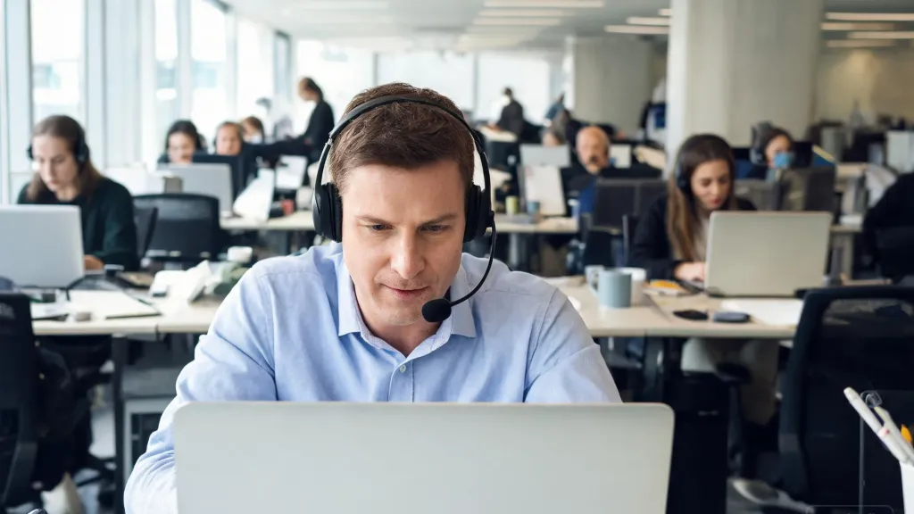 Professional man in an open-plan office wearing a wireless headset, focused on his laptop while colleagues work at neighboring desks