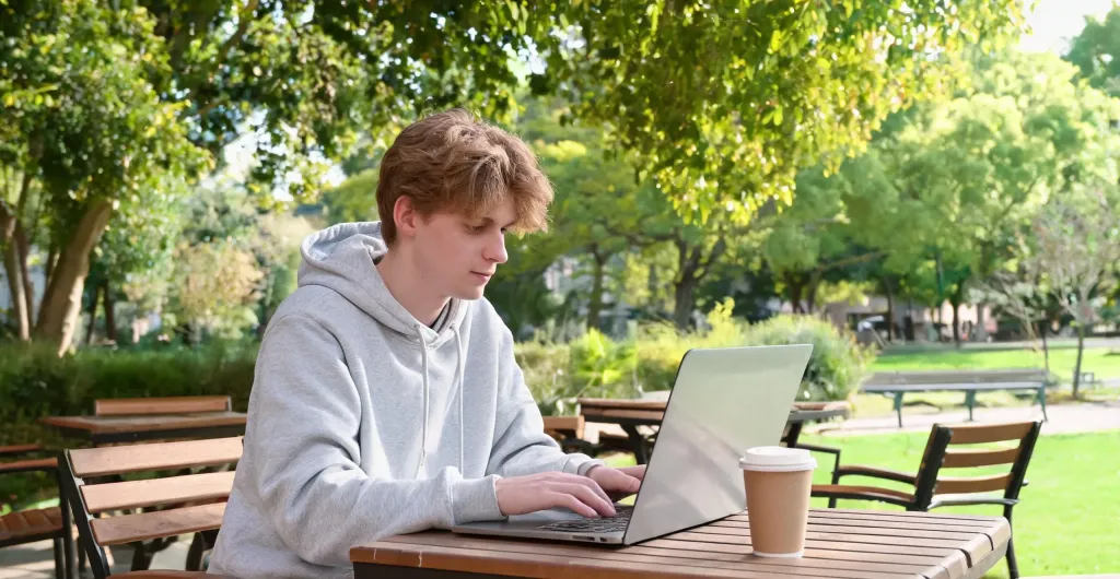 Male university student working at an outdoor campus café table with laptop open, trees and greenery in the background, natural daylight, relaxed atmosphere, lifestyle editorial photography, no text, no logos, clean edges, no borders, no black bars, full bleed composition, edge-to-edge content, no text, no logos, no watermarks, no signatures, no UI badges, no stamps