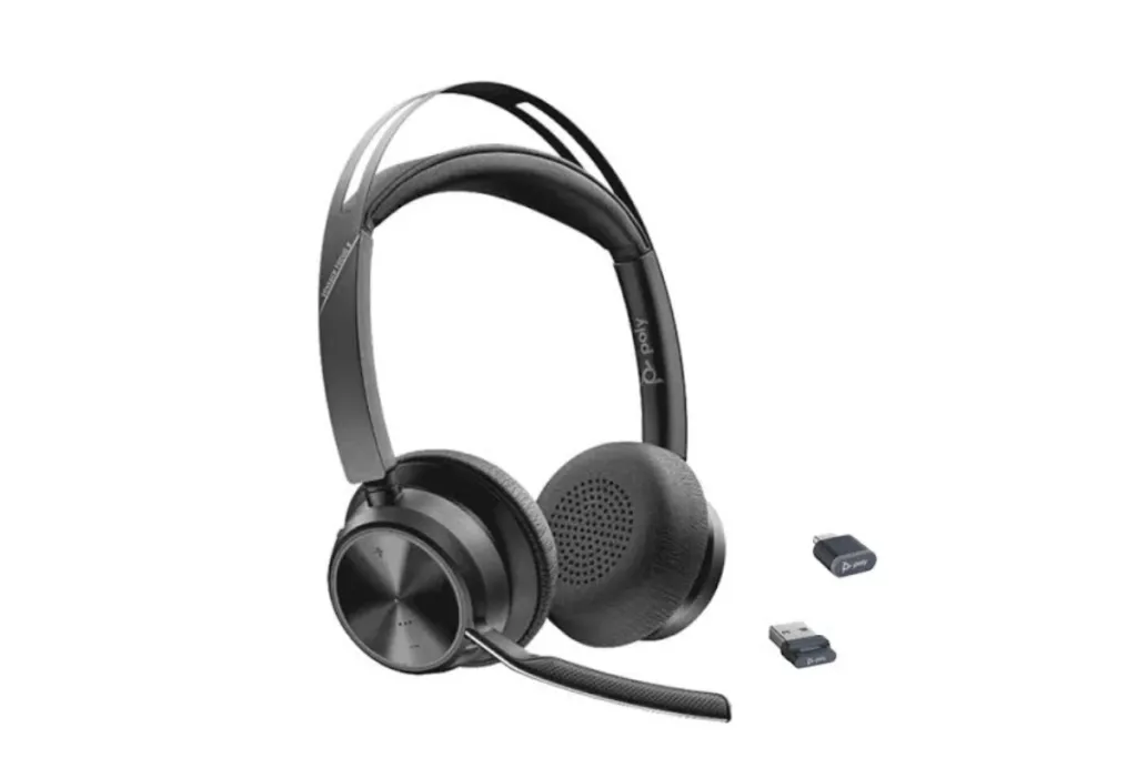 A 3/4 view of a Poly wireless headset with its charging stand and two USB adapters on a white background.