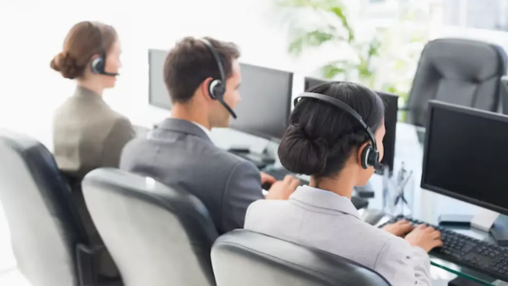 A call center agent working at a desk, speaking on the phone in a busy office environment.