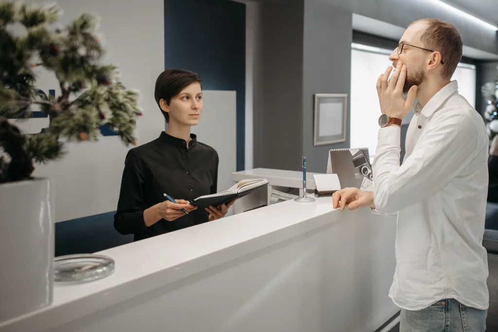 A female receptionist holding a notebook while assisting a male client at a clinic front desk.