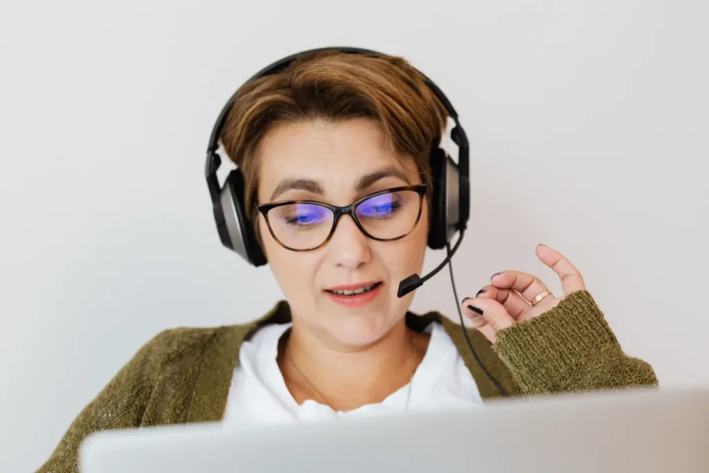 A smiling woman with short hair, wearing glasses and a headset, gestures with her hand during a video call on her laptop.