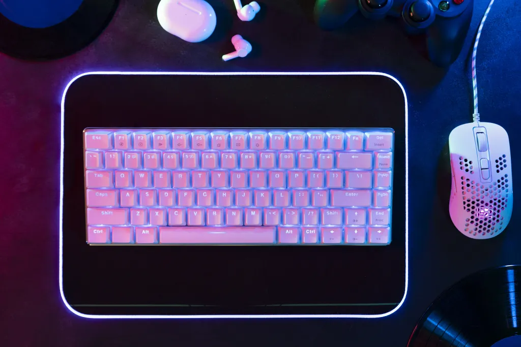 Top view of a bright white mechanical keyboard on an illuminated desk mat next to true wireless earbuds, representing hardware that pairs with the best noise cancelling headphones for coding.