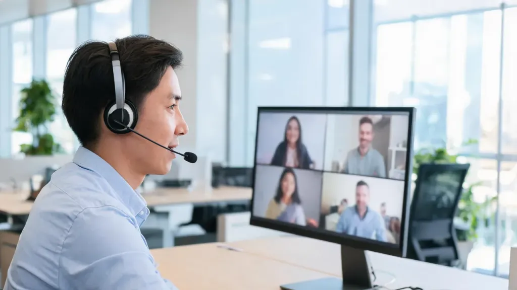 A professional in an office taking part in a video meeting with a headset, colleagues visible on the monitor