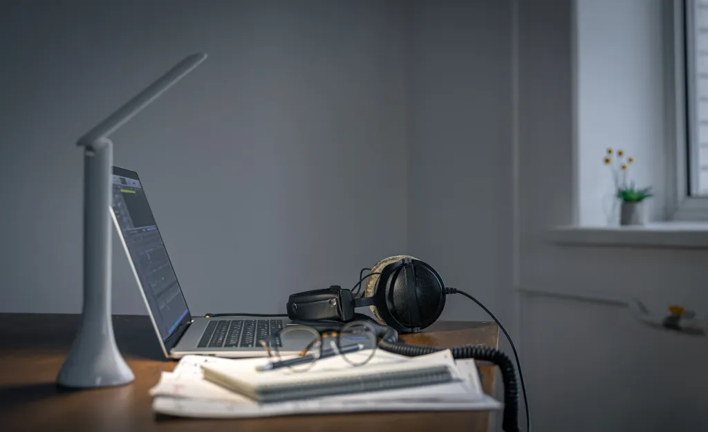 An evening workstation featuring a laptop, reading lamp, and a wireless headphone with noise cancelling microphone resting on some paperwork.