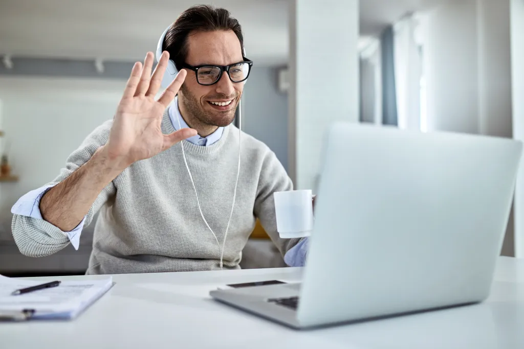 A happy businessman wearing headphones and waving during a video meeting, demonstrating how to seamlessly call phone from pc.
