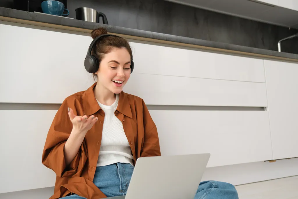 A young female freelancer sitting on the floor using her laptop while wearing a comfortable wireless headphone with noise cancelling microphone for a video call.